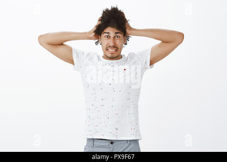 Poor man got stuck in serious problems, feeling desperate and anxious. Worried cute hispanic male model with dark skin, clenching teeth and staring with fright at camera, touching hair nervously Stock Photo