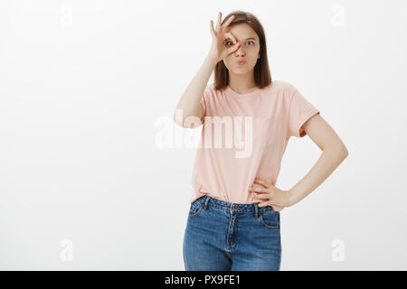 Lifestyle and carefree emotions concept. Good-looking funny woman in pink t-shirt and jeans, holding hand on hip and making circle with eye, showing ok or okay gesture over eye, pouting, having fun Stock Photo