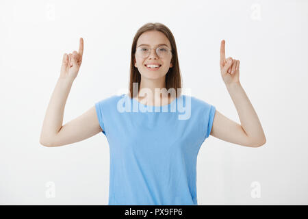 Positive caucasian fair-haired female student in glasses, raising index fingers and pointing up while smiling broadly, sharing great and friendly attitude, being satisfied and happy over grey wall Stock Photo