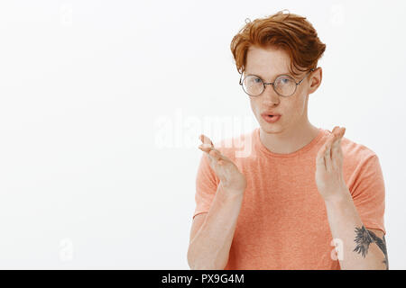 What the hell are you saying. Portrait of bothered intense and annoyed handsome young male with ginger hair and tattoos, folding lips and gesturing with palm while trying to prove man is dumb Stock Photo