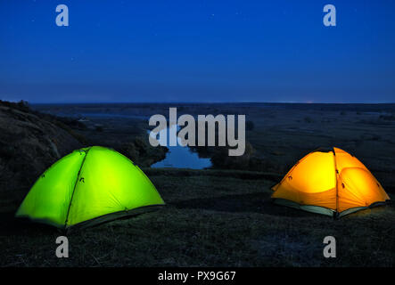 Lighted from the inside orange and green tents on a hill above the river. Night landscape. The concept of freedom, privacy and travel. Stock Photo