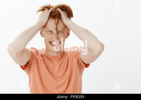Hair scratching badly, need wash it. Portrait of displeased troubled and bothered charming redhead male with freckles in orange t-shirt, pulling hair from head and clenching teeth, being puzzled Stock Photo