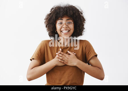 African American girl appreciate friend support being touched with kind words holding hands on heart smiling broadly with grateful happy expression standing pleased and joyful over gray background Stock Photo