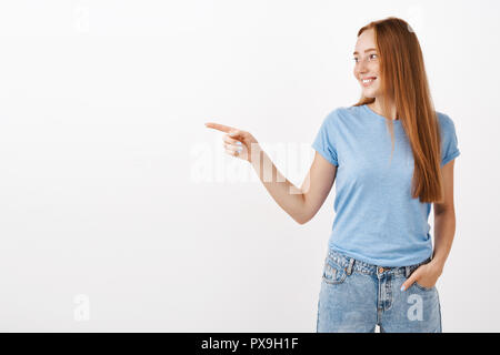 Enjoying watching kids playing in yard. Portrait of cute and tender feminine redhead woman with freckles in blue t-shirt holding hand in pocket pointing and gazing left with delighted happy look Stock Photo