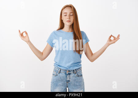Girl finding peace in meditation. Rlieved redhead female with freckles smiling calm closing eyes and standing in lotus pose with fingers in zen gesture concentrating and focusing on inner self Stock Photo