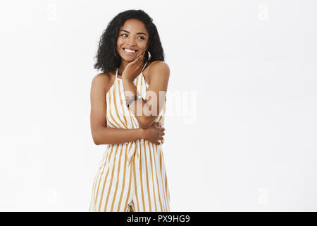 Portrait of charming elegant dark-skinned female enjoying awesome party wearing cute yellow striped overalls touching face gently while chuckling reacing on compliment blushing feeling touched Stock Photo