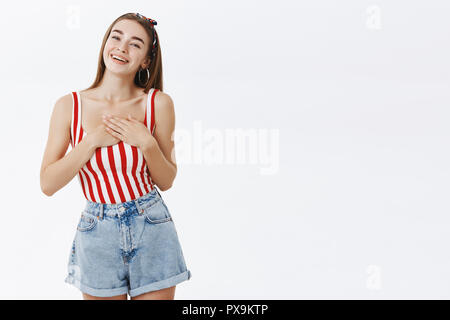Stylish arrogant caucasian female being delighted and thankful receiving compliment standing flirty in pinup striped top, headband tilting head holding palms on heart feeling touched with nice words Stock Photo