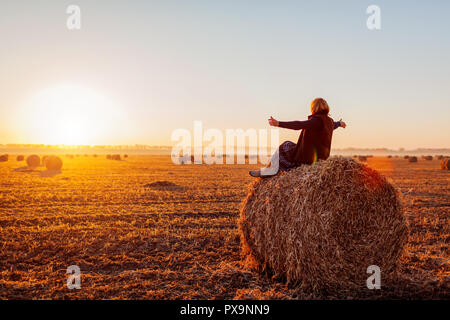 Happy middle-aged woman sitting on haystack in autumn field and feeling free with arms opened. Relaxing and admiring nature at sunset Stock Photo