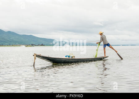 Travel local young Burmese male fisherman wearing checked shirt, using stick and net to fish, balancing on one foot on boat, Inle Lake Myanmar, Burma