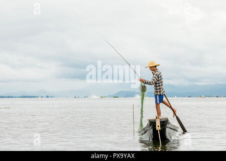 Travel local young Burmese male fisherman wearing checked shirt, using stick and net to fish, balancing on one foot on boat, Inle Lake Myanmar, Burma