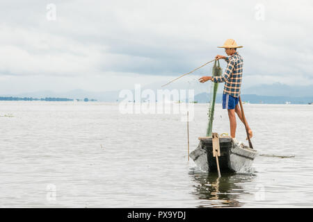 Travel local young Burmese male fisherman wearing checked shirt, using stick and net to fish, balancing on one foot on boat, Inle Lake Myanmar, Burma