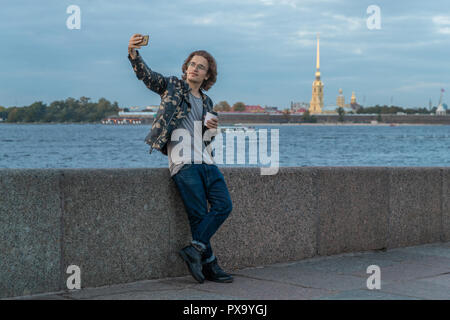 The young man waiting someone and does selfie, drinks coffee, he dressed in a military jacket and jeans, Peter and Paul fortress, cathedral Stock Photo