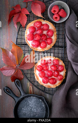 Red raspberry shortbread tarts with vanille custard and glazed fresh raspberries on cooling rack over dark brown texture background. Top view Stock Photo