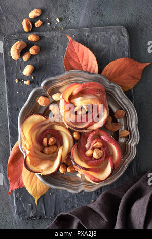Three homemade puff pastries with rose shaped apple slices on metal plate. Top lay on wooden board with Autumn leaves and caramelized nuts. Stock Photo