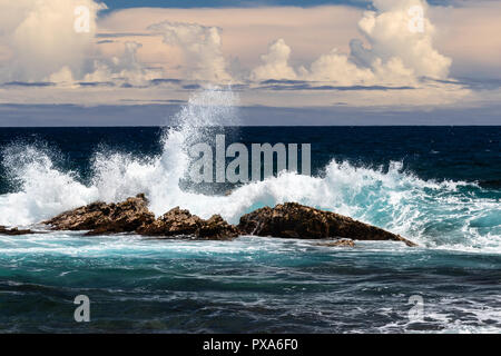 Wave crashing on dark rock; white spray in the air, wave to one side, dark ocean and clouds in background. Black Sand Beach, Punaluu, Hawaii. Stock Photo