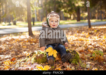 little boy in the pilot cap sitting on the stump, yellow and orange foliage around him. Autumn Stock Photo