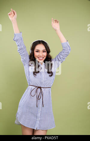 the beautiful young girl listens to music in earphones and dances with the raised hands up against light green background. Stock Photo