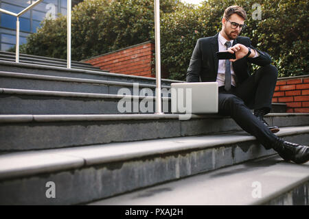 Business professional sitting on steps outdoors with laptop checking time. Young businessman waiting for someone outdoors. Stock Photo