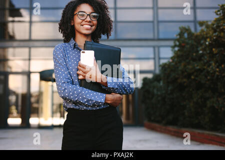 Confident woman in formal clothes holding a smartphone and file looking away smiling. Female executive standing outside office building. Stock Photo