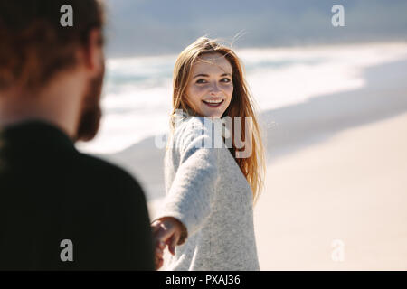 Beautiful young woman holding hand of her boyfriend and walking along the seashore. Couple strolling on the beach holding hands. Stock Photo