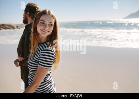 Pretty young woman looking back and smiling while walking on the beach with her boyfriend. Young couple on the sea shore. Stock Photo