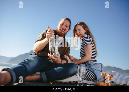 Beautiful couple on picnic at the sea shore opening a champagne bottle. Man and woman having picnic at the beach with food and drinks. Stock Photo