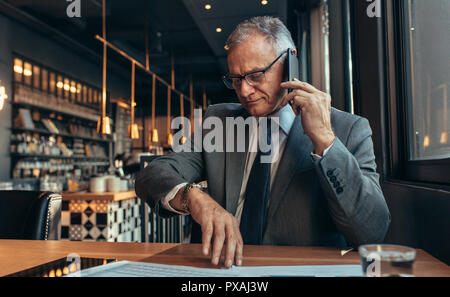 Senior businessman sitting at cafe making a phone call and checking time. Business professional waiting for someone at coffee shop talking over phone. Stock Photo