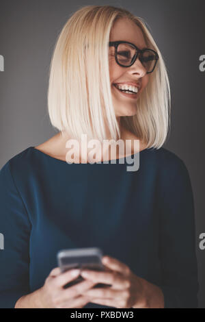 Young businesswoman reading a text message on her cellphone and laughing while standing against a gray background Stock Photo