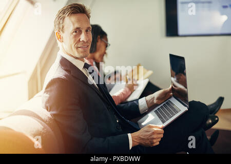 Smiling young businessman working on a laptop while sitting with a group of coworkers during an office presentation Stock Photo