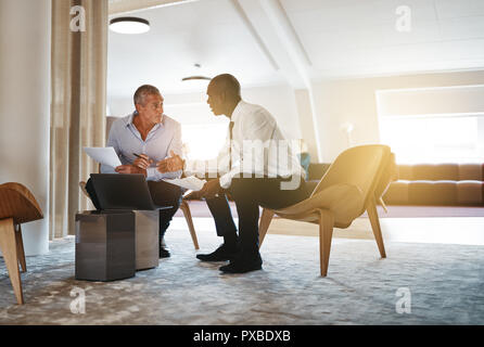 Two diverse businessmen discussing work together and using a laptop while sitting in a modern office Stock Photo