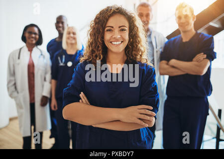 Young female doctor smiling while standing in a hospital corridor with a diverse group of staff in the background Stock Photo