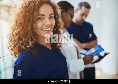 Smiling young female doctor standing in a hospital corridor with two medical colleagues talking in the background Stock Photo