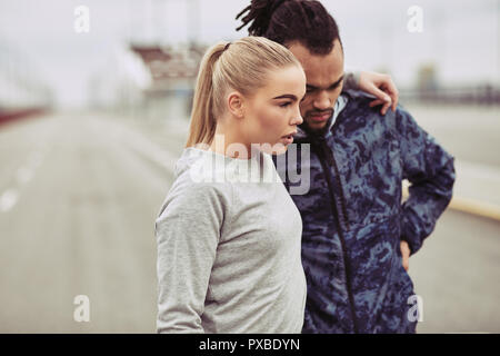 Diverse young couple standing together on a road looking focused while preparing for a run on an overcast day Stock Photo