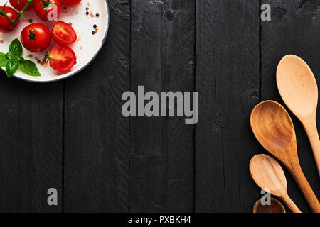 Various wooden spoons and tomatoes in a plate with Himalayan salt, pepper and basil on black wooden table. Top view with copy space for menu or recipe Stock Photo