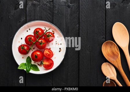 Various wooden spoons and tomatoes in a plate with Himalayan salt, pepper and basil on black wooden table. Top view with copy space for menu or recipe Stock Photo