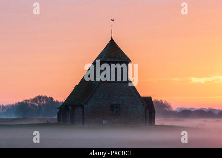 Sunrise and orange cloudy sky over the lonely Thomas a Becket church at Fairfield on Romney Marsh. Fields covered in mist, laying the landscape. Some sheep and cows in the mist. Stock Photo
