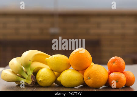 Still life with oranges, lemons, bananas and tangerines placed together in groups, on a background and surface of brown tones Stock Photo