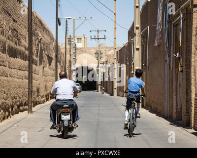 YAZD, IRAN - AUGUST 19, 2016: Old man and young boy, riding a motocycle scooter and a bicycle in the streets of the old Yazd, the main city of central Stock Photo