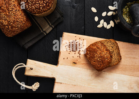 Cutting board with slices of bread, linseed and various crusty whole wheat bread, pumpkin seeds and mortar on black wooden table. Top view. Stock Photo