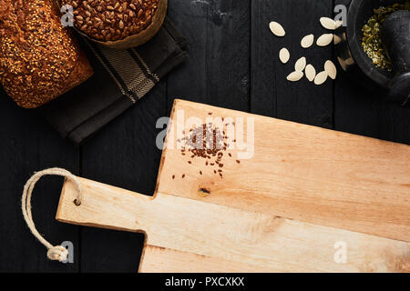 Chopping board with linseed and various crusty whole wheat bread, pumpkin seeds and mortar on black wooden table. Top view with copy space for text, m Stock Photo