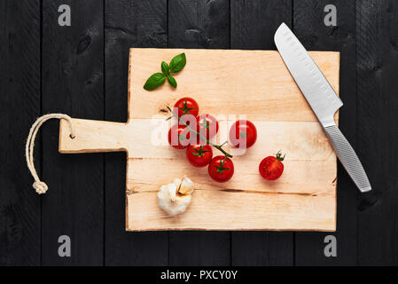 Top view of chopping board with stainless steel vegetable knife, tomato garlic and basil on black wooden background. Stock Photo