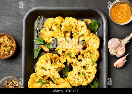 Roasted cauliflower steaks with herbs and spices on baking sheet over black stone background, top view. Vegetarian food. Stock Photo
