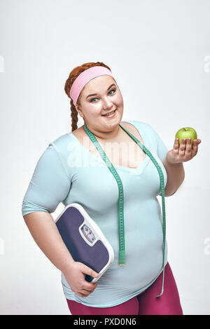 Funny picture of amusing, red haired, chubby woman on white background. Woman holding an apple and scales. Stock Photo
