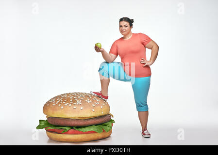 Funny picture of brunette, bearded, plump woman on white background. Woman wearing sportswear. She put her leg to a huge sandwich Stock Photo