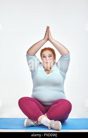 Funny picture of amusing, red haired, chubby woman on white background. Woman trains on the mat. Stock Photo