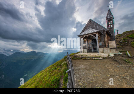 Wooden church at the summit of this mountain in the Italian Alps. Sunbeam, sunray filtering through the clouds. Holy light from the sky. Bell tower. Stock Photo