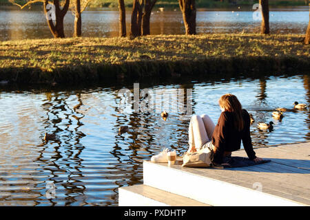 Back view of long hair brunette girl sitting on the pier near lake at sunset time Stock Photo
