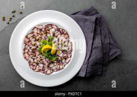 OCTOPUS CARPACCIO. Seafood Raw octopus slices with olive oil, lemon and capers on white plate. Top view. Gray stone background. Stock Photo