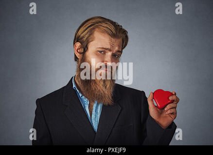 Sad unhappy skeptical hipster businessman in black jacket, blue shirt standing and holding red big heart shaped gift box looking at camera with cry, b Stock Photo