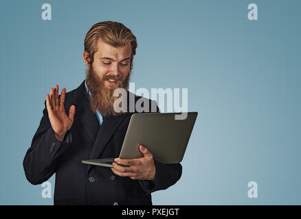 Hi there. Bearded hipster business man having a video call with a laptop greeting someone isolated on blue background with copy space. Multicultural m Stock Photo